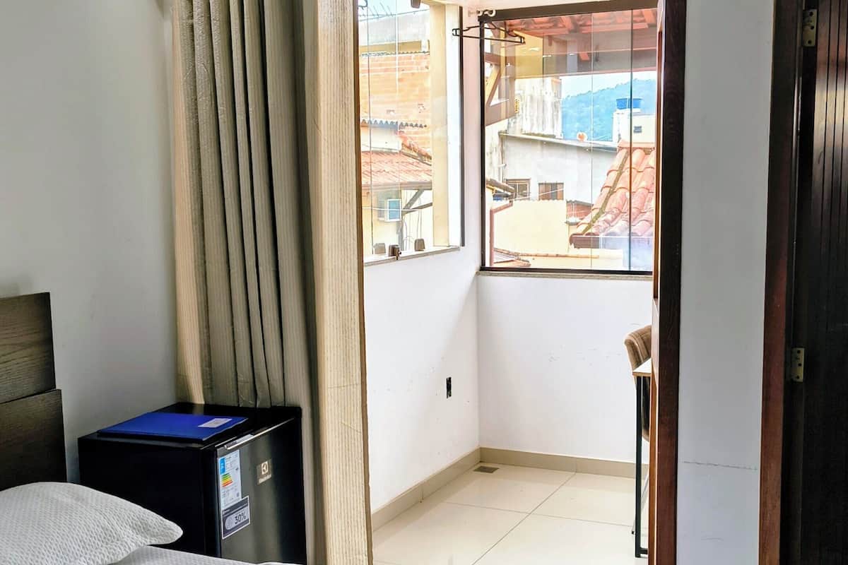 A hotel room with a bed, a desk, an air conditioning unit, a window with curtains, and a view of the outside.