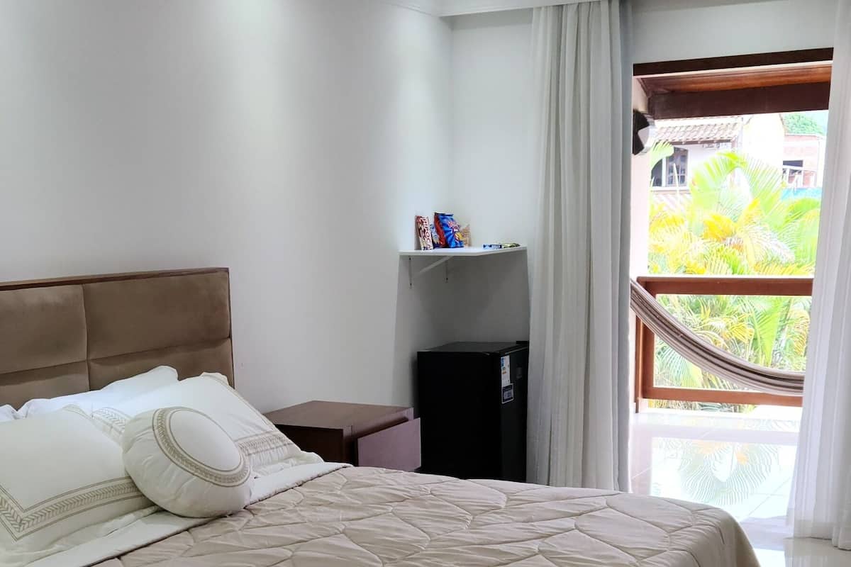 A modern hotel room with a large bed, a bedside table, a small cabinet, and a view of greenery outside.