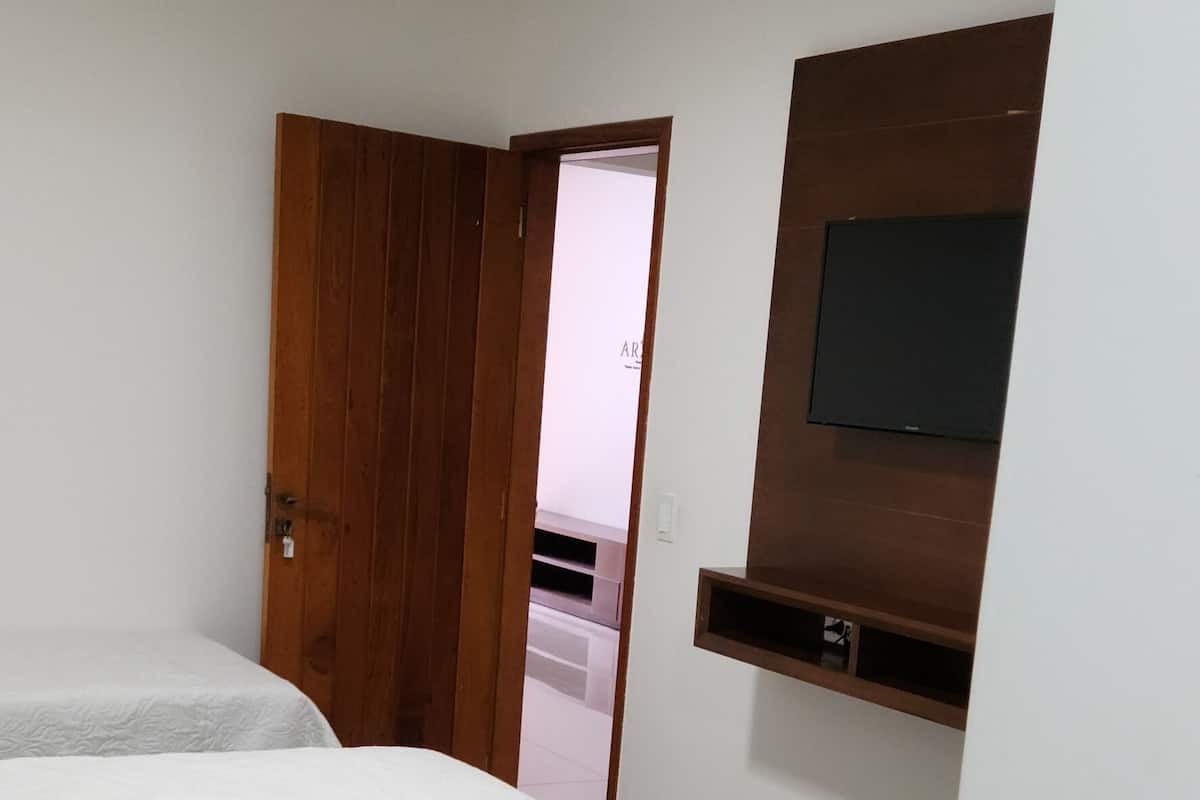 A bedroom with a bed, a wooden door, a TV mounted on the wall, and a ceiling light.