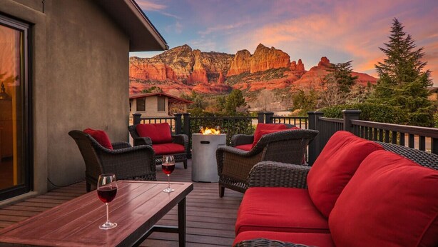 A balcony with wicker furniture, a table with a glass of red wine, and a view of red rock formations. - Walk to Uptown + Hot Tub, Firepit, Epic Views (Sedona)
