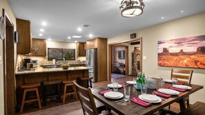 A modern kitchen with wooden cabinets, a central island, and a dining area set for six with a large painting on the wall. - Walk to Uptown + Hot Tub, Firepit, Epic Views (Sedona)
