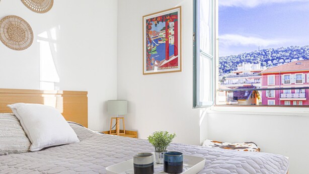 A bedroom with a bed, a window with a city view, a framed picture on the wall, and a tray with two cups on the bed. - Le Palais du Vieux Port by Interhome (Nice)