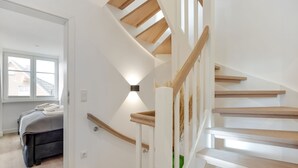 Moderni portaikko, jossa on puiset askelmat ja kaiteet, johtaa makuuhuoneeseen, jossa on sänky ja ikkuna.