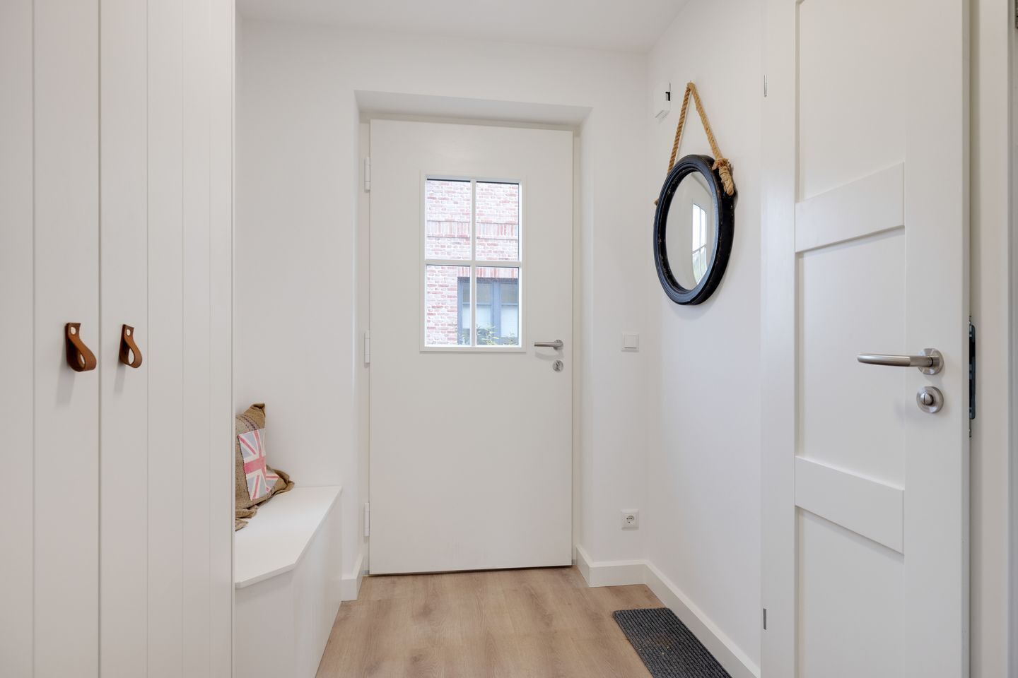 Un couloir étroit avec une porte blanche, une petite fenêtre et un miroir accroché au mur.