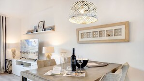A dining area with a wooden table, chairs, a wine bottle, and glasses. There is a television on a white cabinet, a lamp, and a framed picture on the wall. - Thilo - "Charming vacation home on Sylt: relaxation and recreation await you!" (Sylt / OT Tinnum)