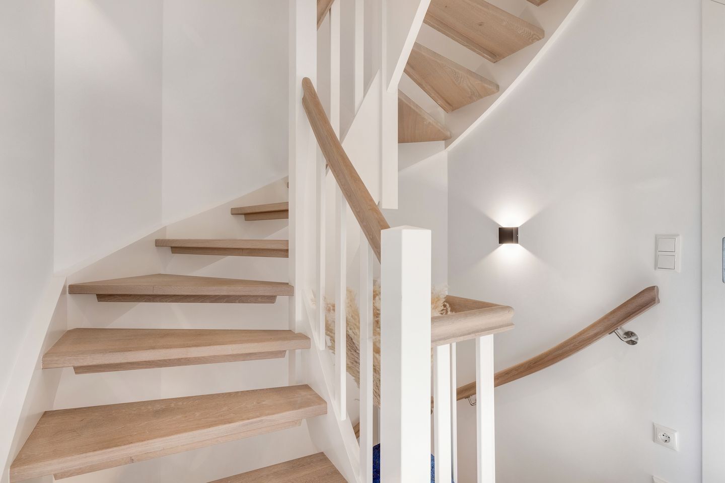 A modern staircase with wooden steps and white railings, leading to a landing with a wall-mounted light fixture.