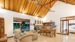 A spacious living area with a dining table, wicker furniture, a kitchen, and a large window with a view of palm trees. - Villa Hijau, Penestanan, Ubud (Ubud, Gianyar Regency)