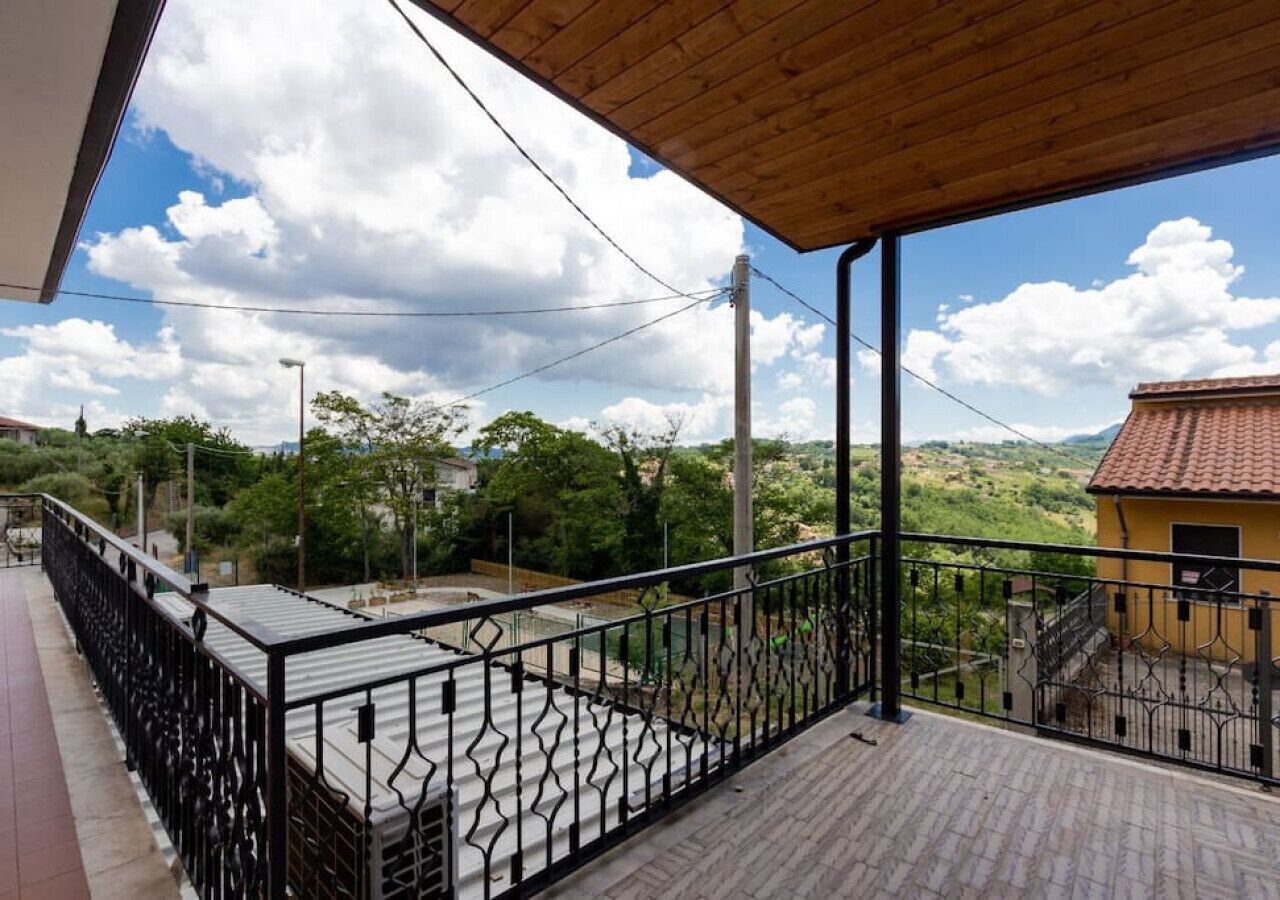 Un balcone con pavimento in legno e ringhiera in metallo, con vista su un paesaggio verde e un edificio giallo.