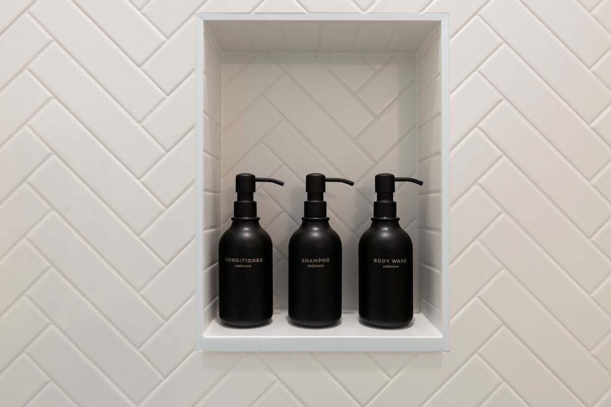 Ein weißes Regal mit drei schwarzen Flaschen, beschriftet mit Conditioner, Shampoo und Duschgel.