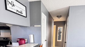 Une cuisine moderne aux tons gris et blancs, un coin bar avec des tabourets, et un couloir menant à une chambre.