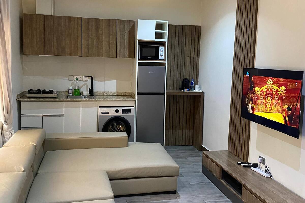 A modern living room with a sectional sofa, a flat-screen TV mounted on the wall, and a kitchen area with a microwave and refrigerator.