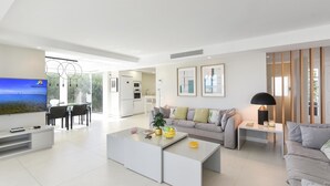 A modern living room with a flat-screen TV, a sofa, a coffee table, and a dining area with chairs.