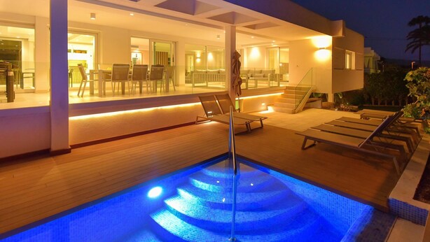 Un espace piscine moderne avec un élément décoratif aquatique illuminé de lumière bleue, un transat et un coin repas doté de murs en verre.