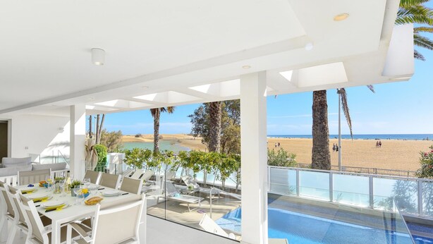 A modern outdoor dining area with a pool, overlooking a beach and desert landscape.