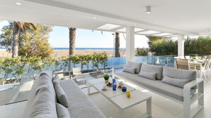 A modern outdoor lounge area with a sofa, chairs, and a dining table, overlooking a beach and the sea.