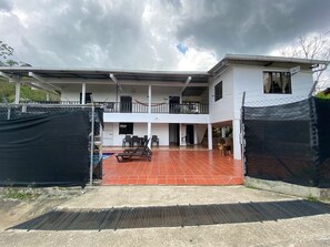 Two-story building with a flat roof and balcony, featuring a patio area and pool in the background.