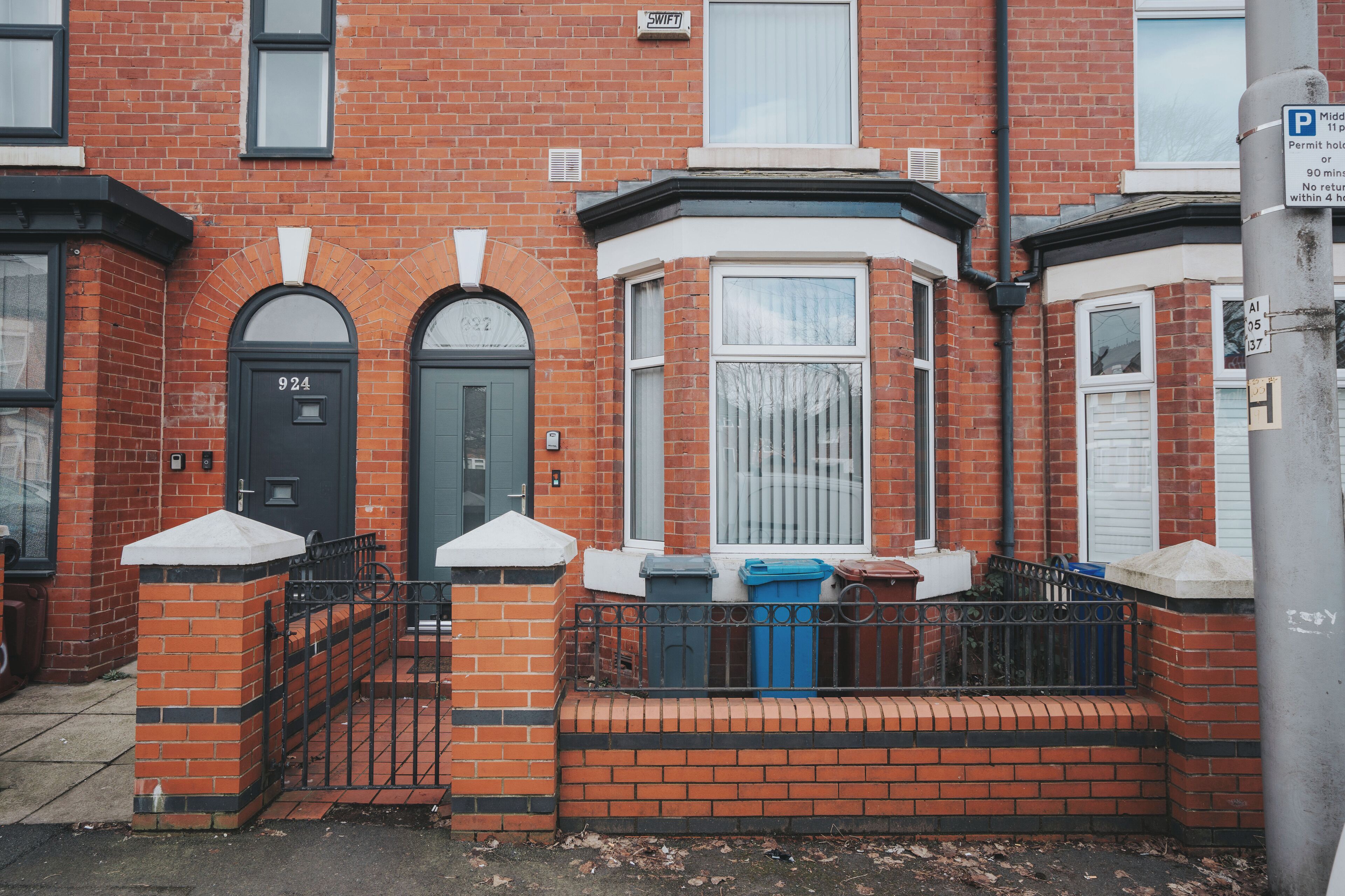 A red brick house with a black front door, a bay window, and a small porch area with trash bins.