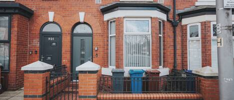 A red brick house with a black front door, a bay window, and a small porch area with trash bins.
