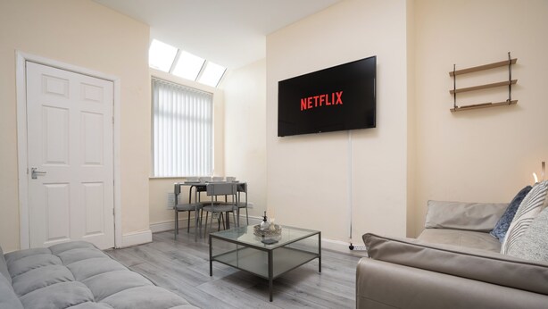 Ein modernes Wohnzimmer mit Sofa, Glastisch, Wandfernseher und einem Essbereich mit Tisch und Stühlen.