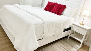 A neatly made bed with white bedding and a red pillow, a bedside table with a lamp, and a white nightstand.