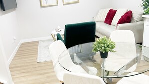 A modern living room with a glass dining table, white chairs, a green sofa, and a red throw pillow.