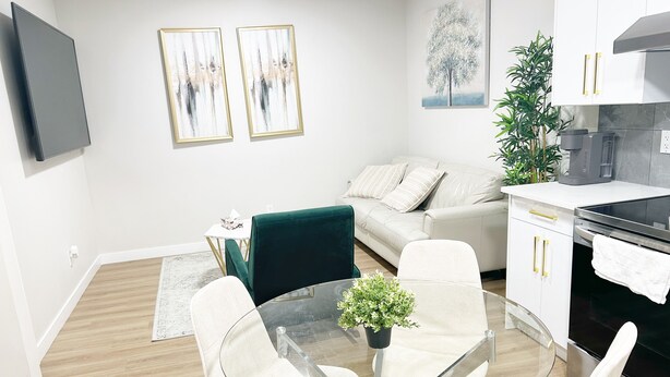 A modern living room with a glass table, white chairs, a green sofa, and a kitchen area with a stove.