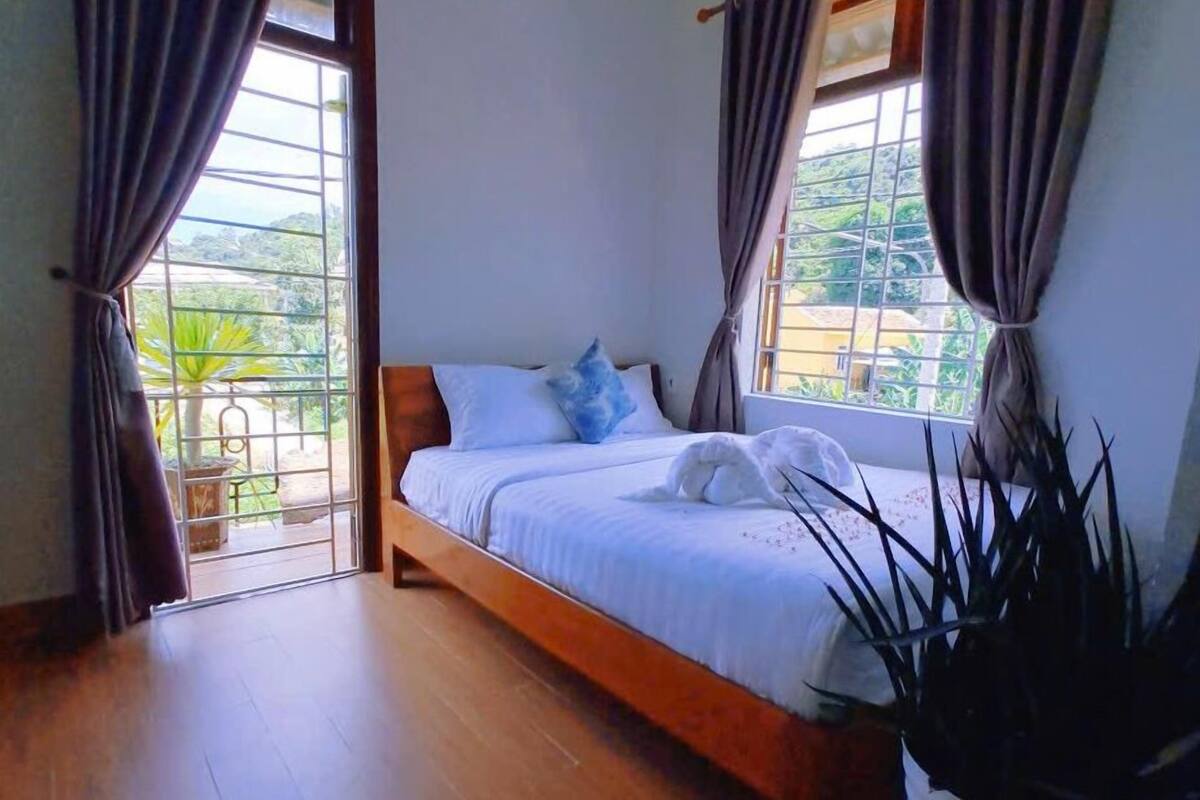 A bedroom with a wooden bed, curtains, a potted plant, and a view of greenery outside.