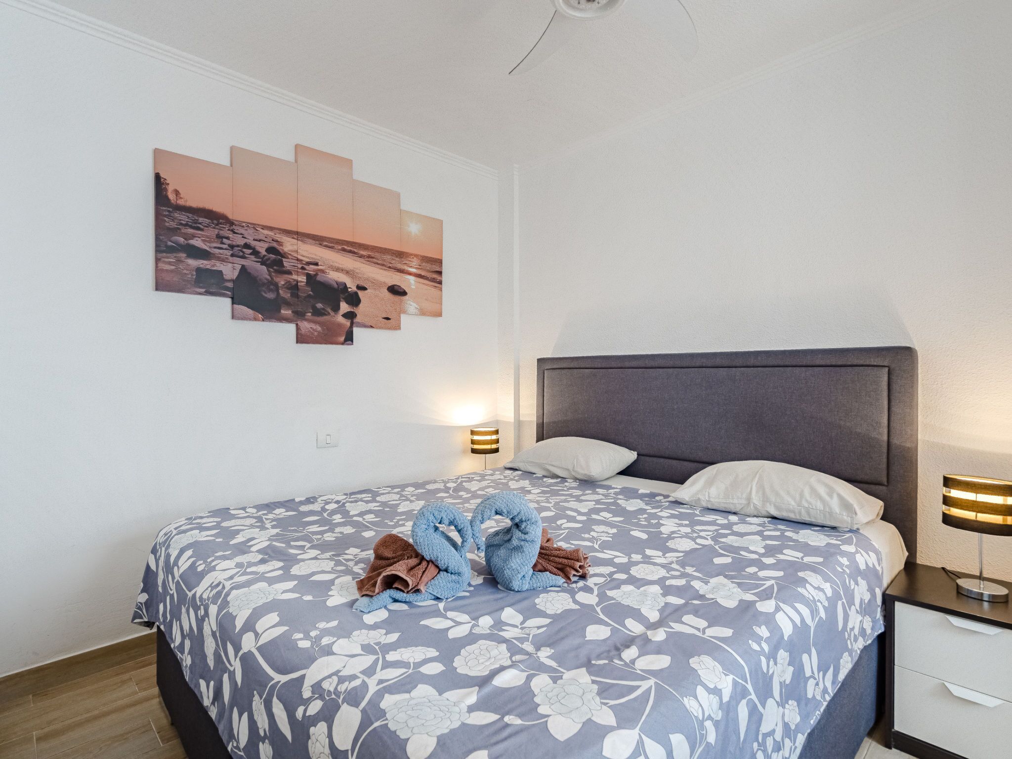 Un lit bien fait, recouvert d’une couvre-lit à motifs floraux, avec une tête de lit et un tableau accroché au mur représentant un coucher de soleil sur des rochers.