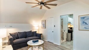Ein Wohnzimmer mit grauem Sofa, einem runden Couchtisch, einem Deckenventilator und einer durchgehenden Tür erkennbaren Küche.