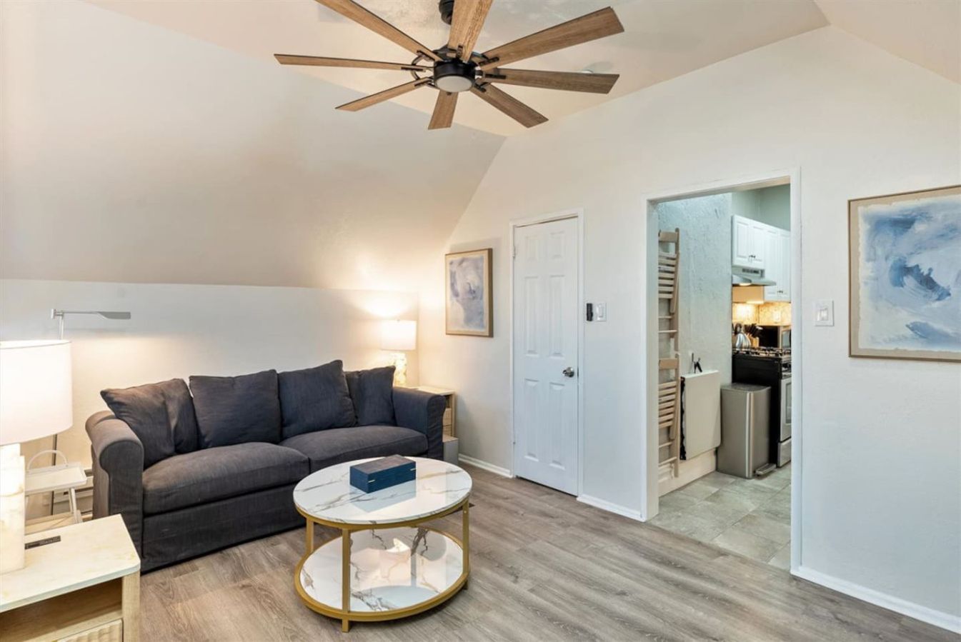 A living room with a grey sofa, a round coffee table, a ceiling fan, and a kitchen visible through an open door.