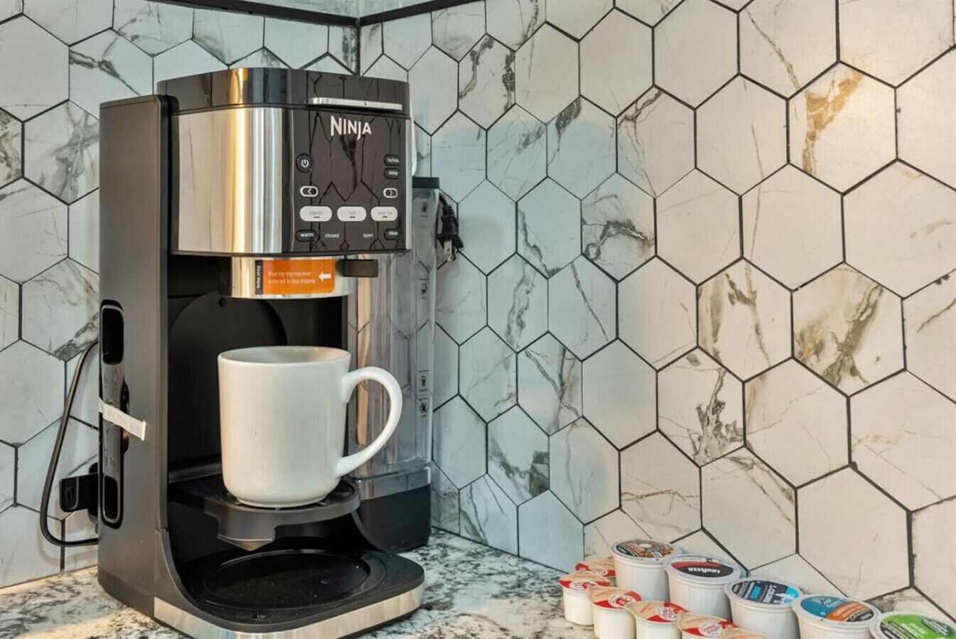 A coffee machine with a cup holder, a hexagonal tiled backsplash, and coffee pods on the counter.