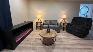 Ein modernes Wohnzimmer mit Kamin, einem grauen Sofa, einem schwarzen Sessel und zwei Beistelltischen mit Lampen.