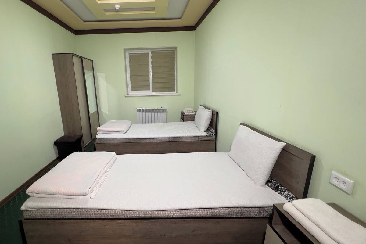 A small, neatly arranged bedroom with two beds, a mirror, a radiator, and a window.