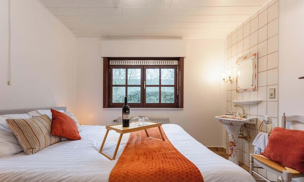 Une chambre d’hôtel avec un lit, une fenêtre, une table de chevet avec une bouteille, une salle de bain avec un lavabo et un miroir, et un petit tabouret.