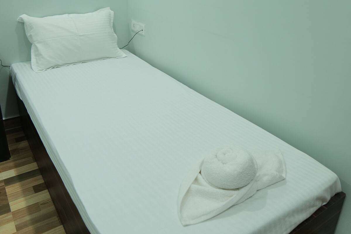 A small, single-bed room with a white bedspread, a white pillow, and a white towel on the bed. The room has light-colored walls and a wooden floor.