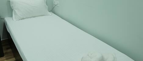Una habitación pequeña con una cama individual, una colcha blanca, una almohada blanca y una toalla blanca sobre la cama. La habitación tiene paredes de color claro y piso de madera.