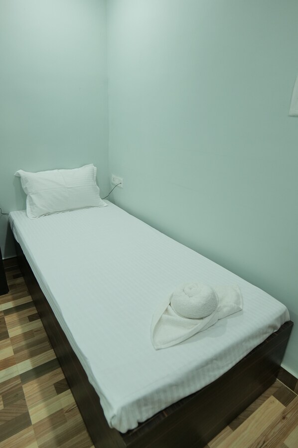 Een kleine kamer met een enkel bed, voorzien van een wit sprei, een wit kussen en een witte handdoek op het bed. De kamer heeft muren in een lichte kleur en een houten vloer.