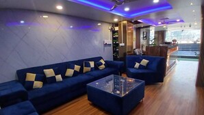 A modern living room with blue sofas, a glass coffee table, wooden flooring, ceiling fans, and a reception area with a counter and plants. - Hotel HK Regency (Guwahati)