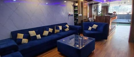 A modern living room with blue sofas, a glass coffee table, wooden flooring, ceiling fans, and a reception area with a counter and plants.
