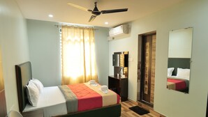A hotel room with a bed, a desk, a chair, a ceiling fan, a window with curtains, a mirror, and a wall-mounted air conditioner. - Hotel HK Regency (Guwahati)