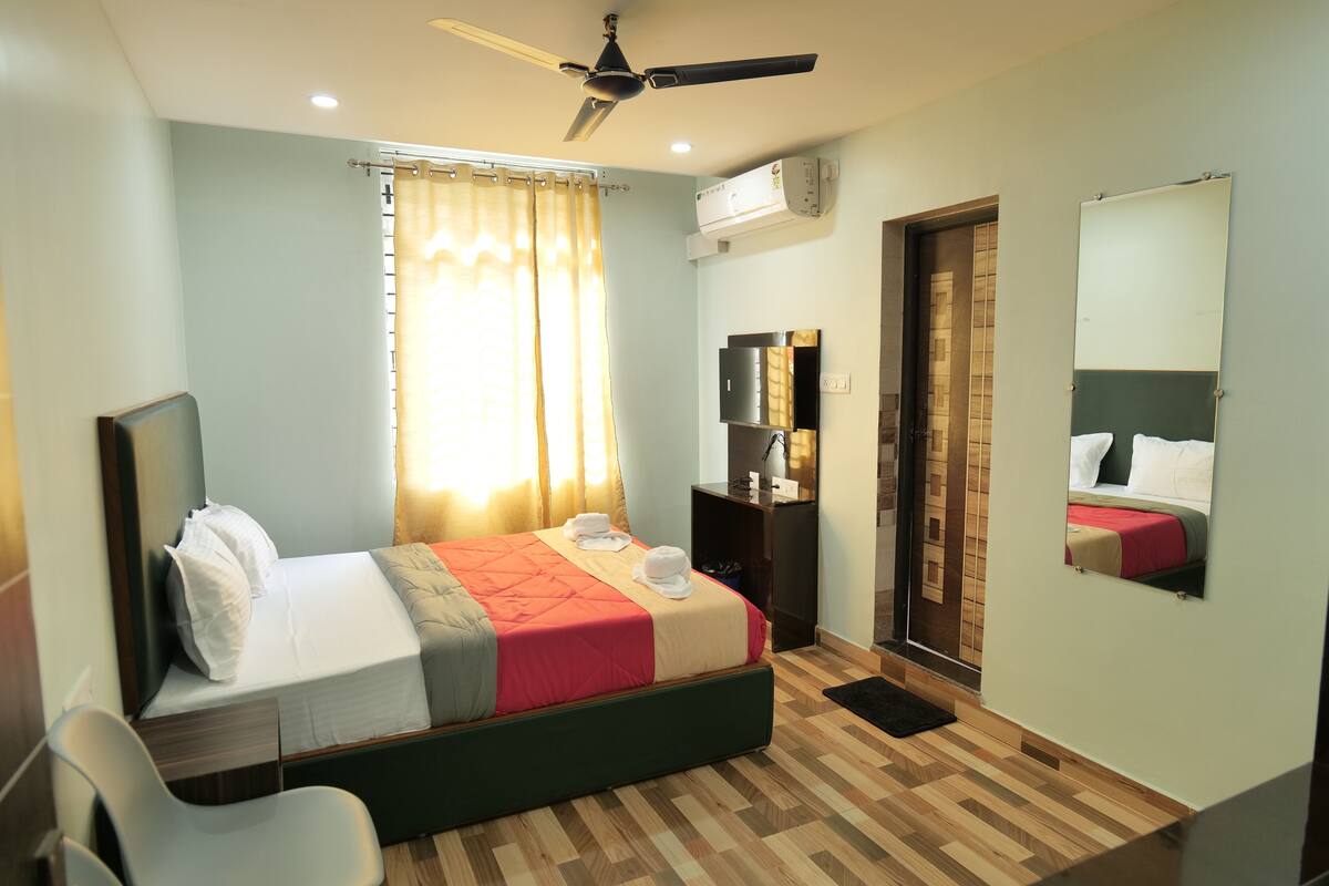A hotel room with a bed, a desk, a chair, a ceiling fan, a window with curtains, a mirror, and a wall-mounted air conditioner.