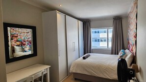Een hotelkamer met een bed, bureau, kledingkast, raam met gordijnen en een kleurrijk schilderij aan de muur.