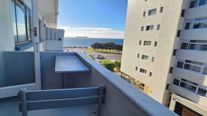 Een balkon met een kleintje tafeltje en een bankje, met uitzicht op een parkeerplaats en een gebouw met veel ramen.