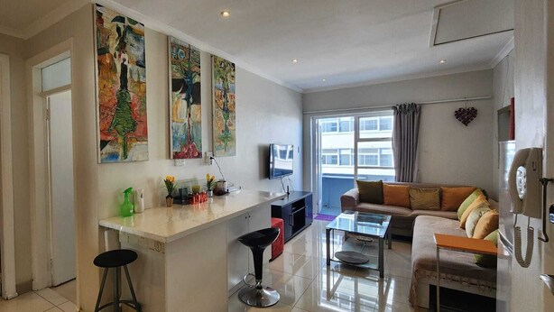 Een moderne woonkamer met een bank, een glazen salontafel en een televisie. Er is een keukenhoek met een bar en barkrukken. De kamer heeft grote ramen en kunstwerken aan de muren.