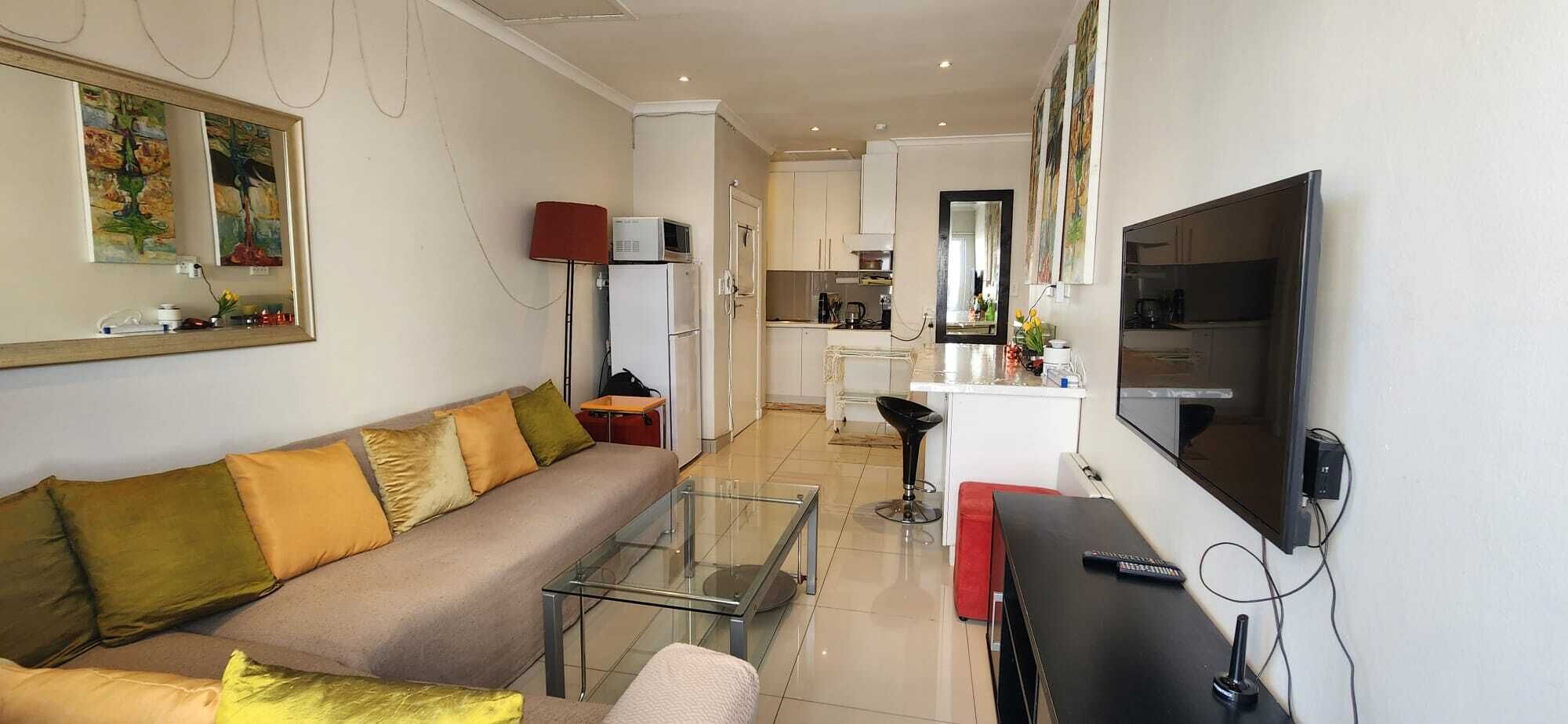 Un salon moderne avec une table basse en verre, un canapé avec des coussins, un téléviseur et un coin cuisine à l’arrière-plan.