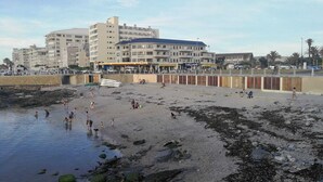 Een strand met mensen die zwemmen en wandelen, een parkeerplaats en gebouwen op de achtergrond.