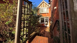 A narrow brick pathway leading to a wooden gate with a window, surrounded by greenery and a brick building with a gabled roof. - Thin End (Seaview)