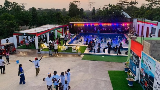 Zona de piscina con gente alrededor, un área de estar cubierta y un edificio con fachada roja y blanca.