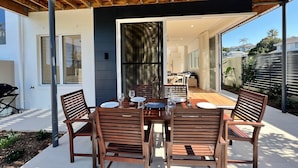 An outdoor patio with a wooden table and chairs, a grill, and a view of the house interior.