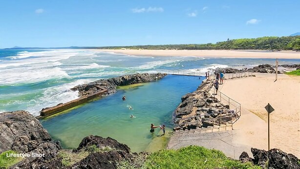 A coastal scene with a clear swimming area surrounded by rocks, a sandy beach, and a view of the ocean.